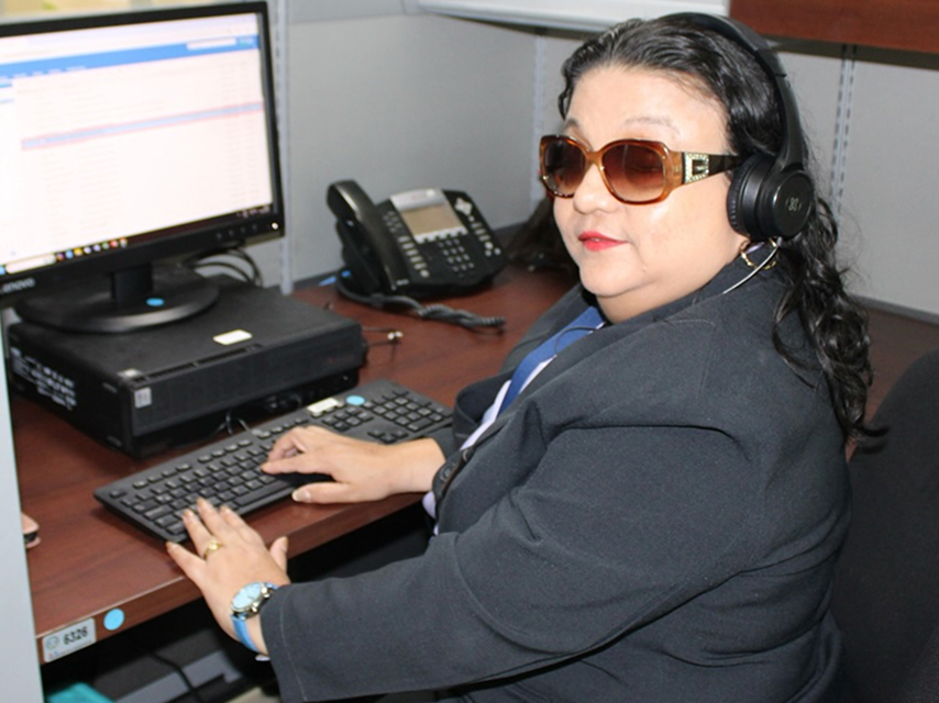 Una mujer de cabello oscuro, usando gafas de sol grandes y audífonos, está sentada en un escritorio de oficina. Tiene sus manos sobre el teclado, frente a un monitor y un teléfono.
