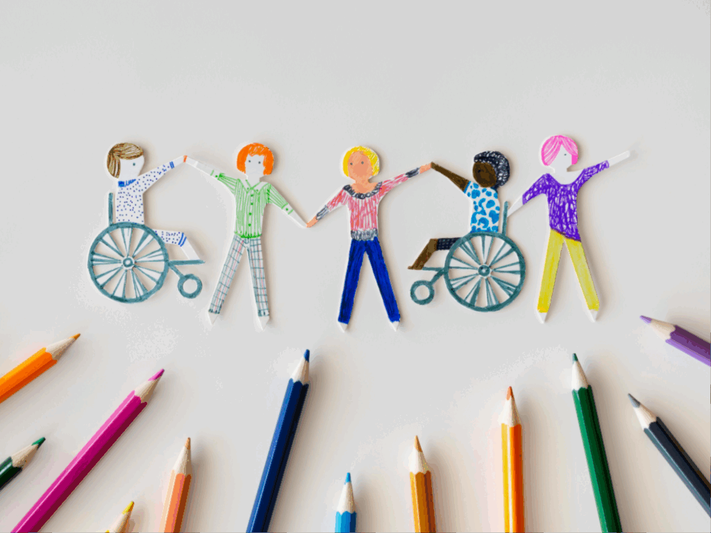 cinco figuras de papel coloreadas a mano, tomadas de las manos. Dos de las figuras están sentadas en sillas de ruedas, y las otras tres están de pie. Todos los personajes tienen atuendos y peinados únicos y coloridos, representando la diversidad. Alrededor de las figuras, hay varios lápices de colores dispersos.